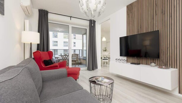 Un salon moderne avec un canapé gris, un fauteuil rouge et un téléviseur fixé sur un mur recouvert de panneaux en bois.