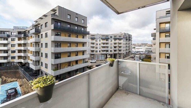 A balcony with a view of multi-story residential buildings, a parking area, and a clear sky.