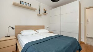 A bedroom with a bed, bedside tables, a nightstand, a wall-mounted shelf with pictures and a plant, and a wardrobe.