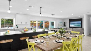 Une cuisine moderne avec un îlot central, un coin repas avec des chaises jaunes et un téléviseur fixé au mur.