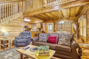 Interior de una cabaña de madera con sofá, sillón, mesa de comedor y zona de cocina.