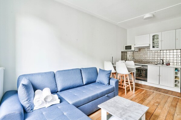 Een moderne woonkamer met een blauwe bank, een klein tafeltje, en een keukenhoek met witte kasten en een magnetron.