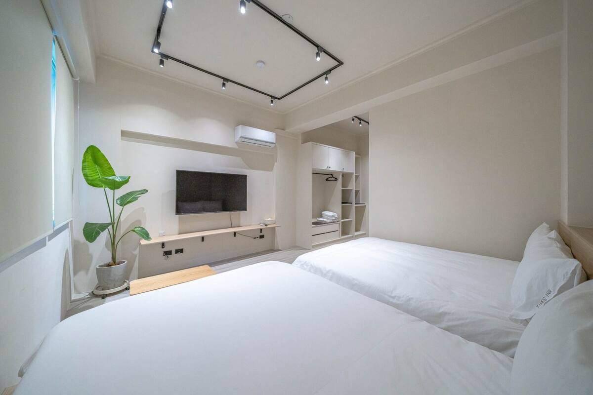 A modern hotel room with two beds, a flat-screen TV, built-in shelving, and a potted plant.