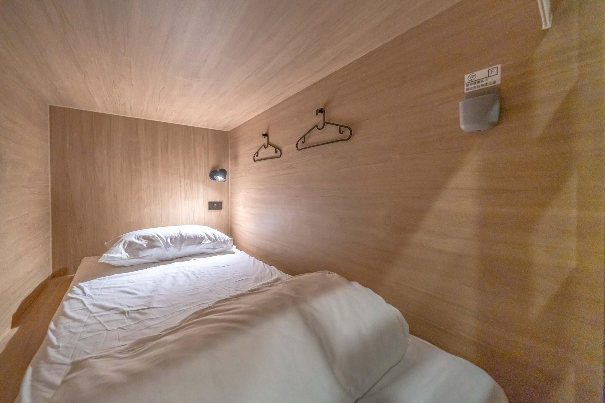 Una habitación con paredes de madera, una cama con ropa de cama blanca, dos perchas fijadas a la pared y una lámpara pequeña también en la pared.