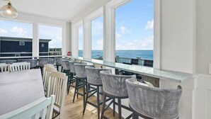 A seaside bar area with high windows, wicker chairs, and a view of the ocean. - Admirals Lodge | Pool & Hot tub | Games | Oceanfront (Oak Island)