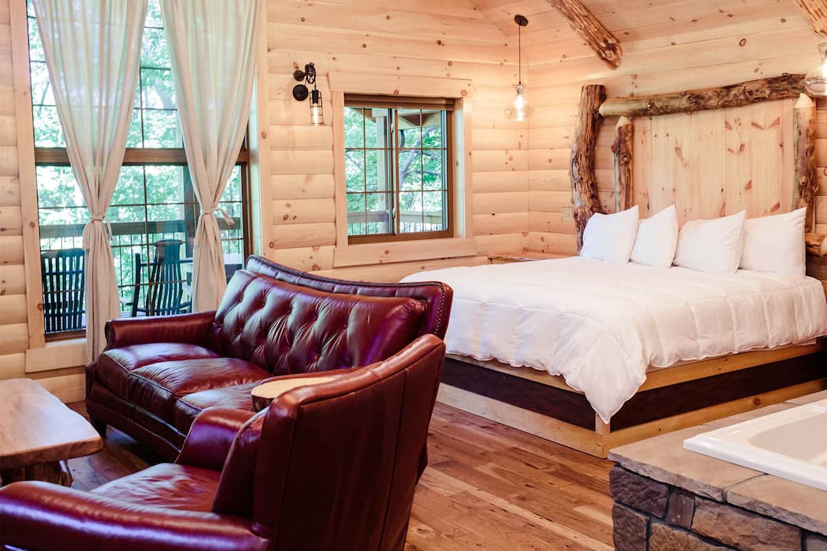 A log cabin bedroom with a large bed, a leather sofa, a wooden floor, and a stone bathtub.