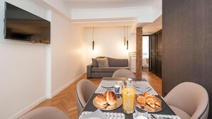 Un comedor con una mesa dispuesta para el desayuno, que incluye croissants y jugo, en un apartamento moderno con un televisor montado en la pared y un sofá al fondo.