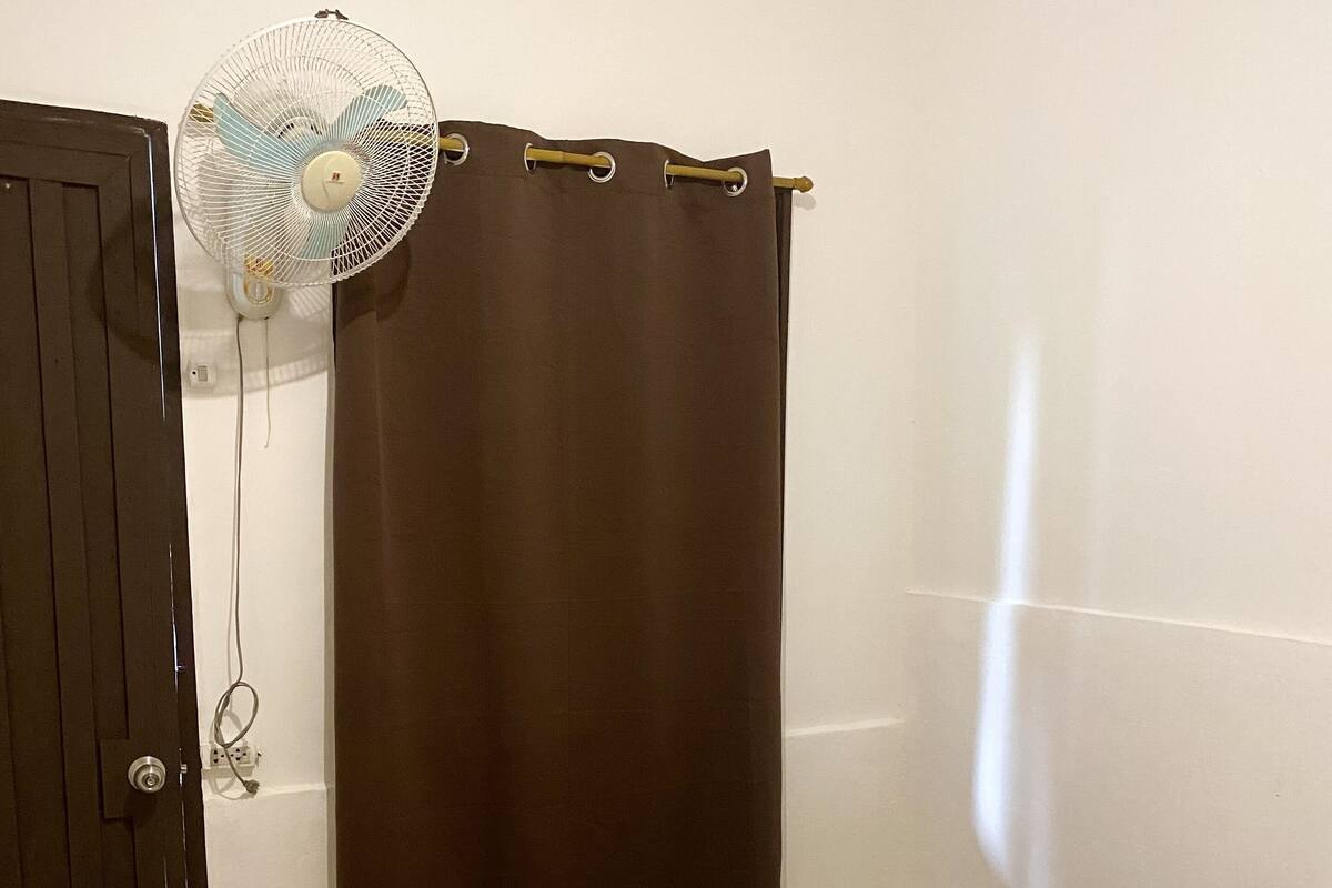 Une pièce avec un lit, un rideau marron, un ventilateur blanc et une porte.