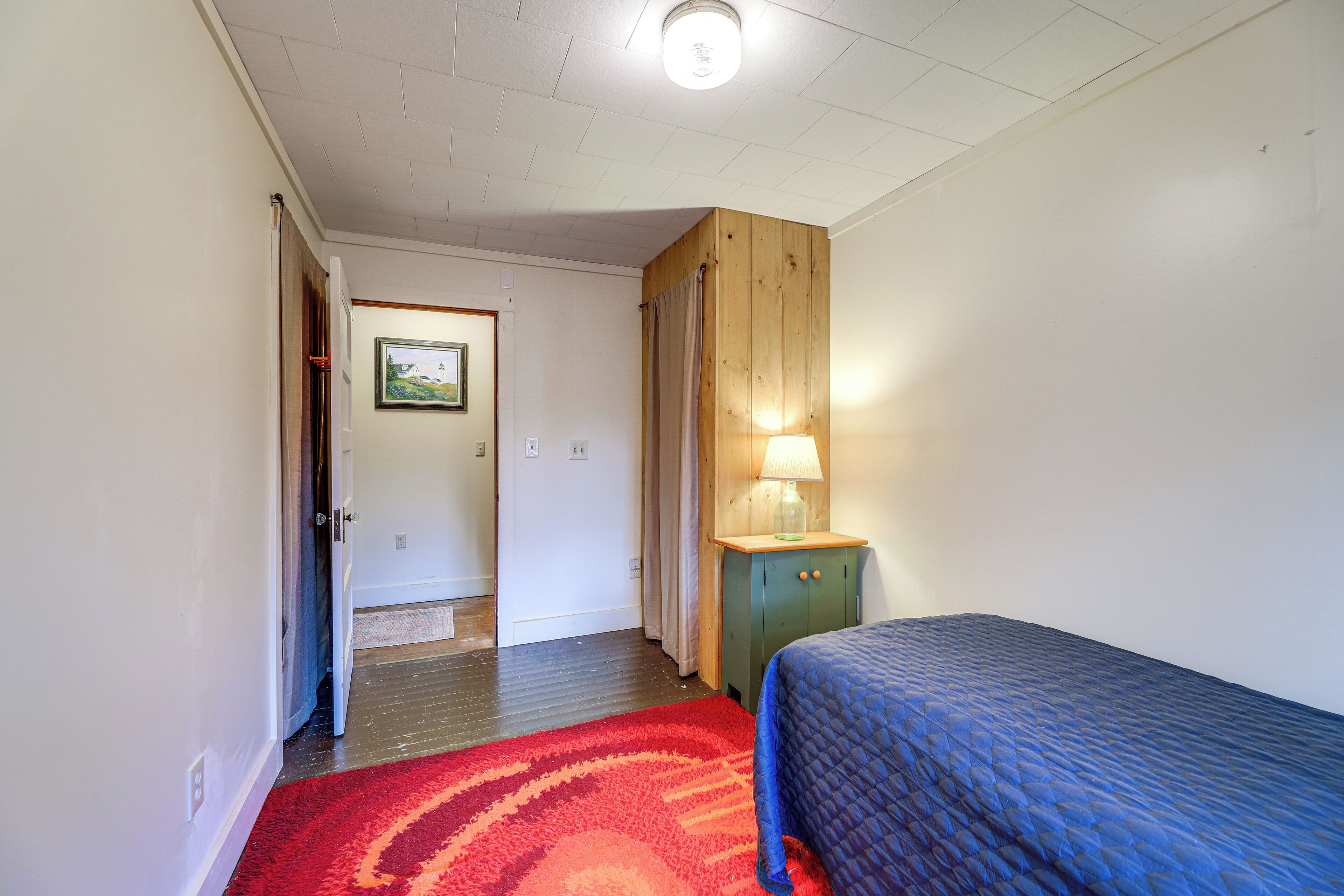 Une chambre à coucher avec un lit, une armoire en bois surmontée d’une lampe, un tapis rouge et une porte donnant sur une autre pièce.