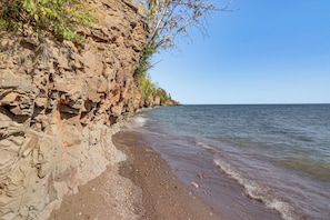 A rocky shoreline with clear water, a sandy beach, and a cliff with sparse vegetation.
