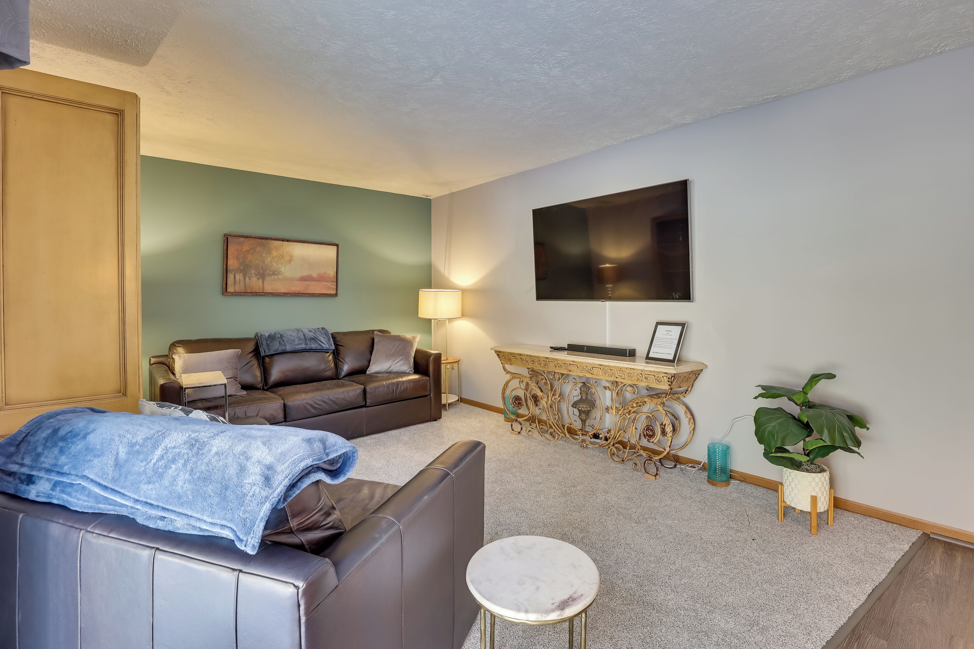 A living room with a sofa, a small round table, a TV mounted on the wall, a decorative console table, and a potted plant.