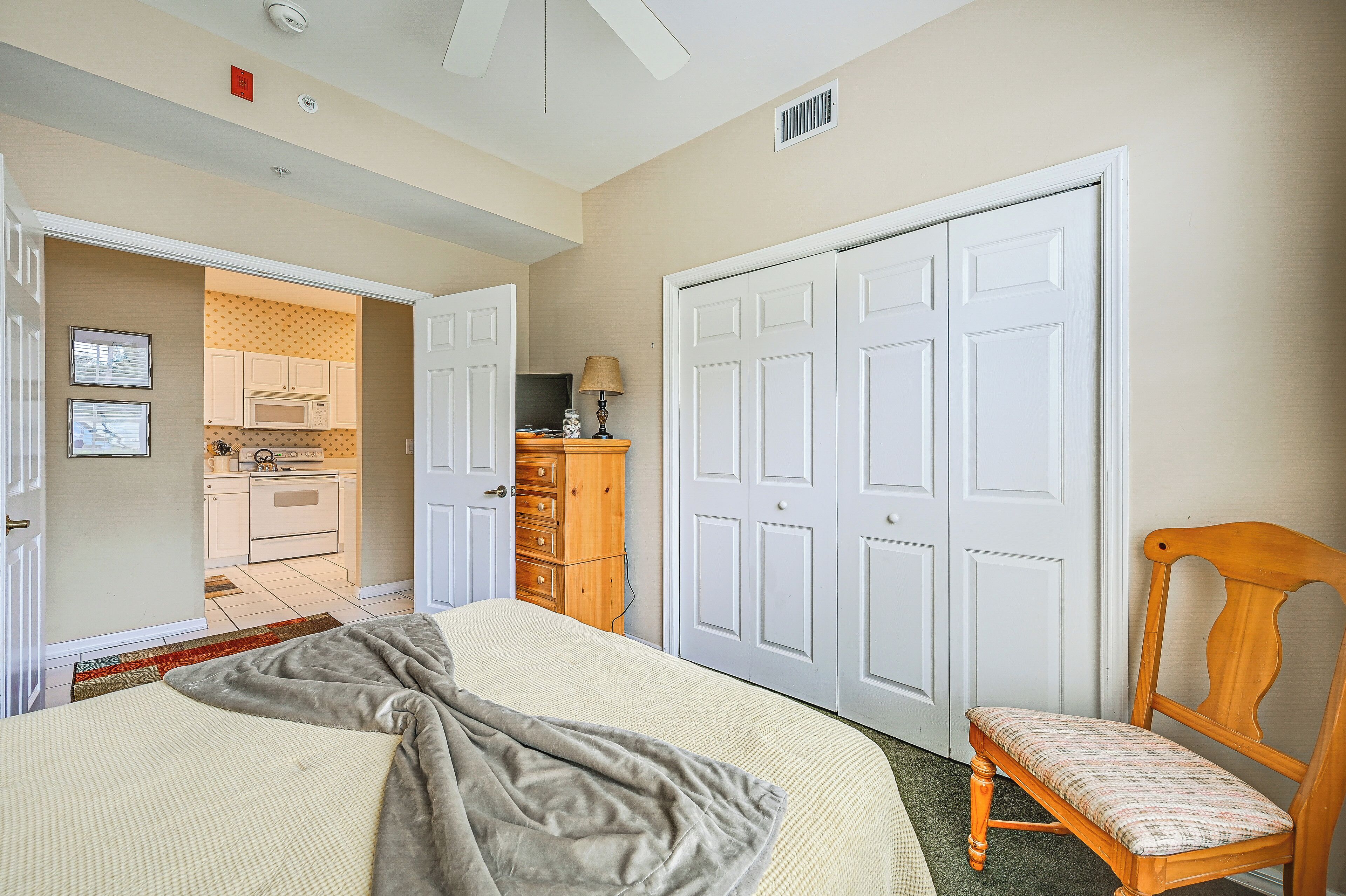 A bedroom with a bed, a wooden chair, a wardrobe, and a kitchen in the background.