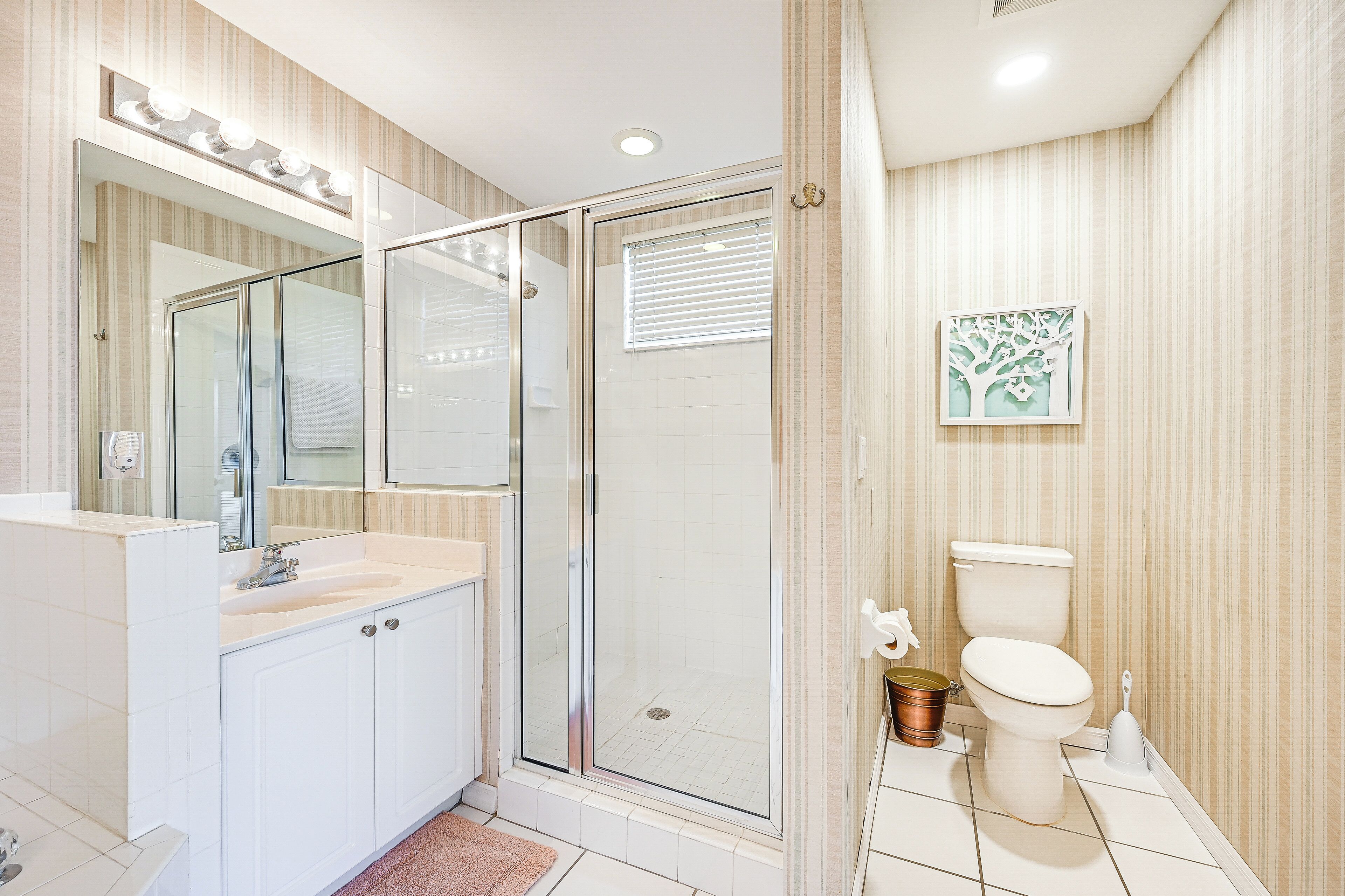 Een moderne badkamer met een glazen douchecabine, een wit wastafeltje met spiegel erboven en een toilet met een decoratief kunstwerk van een boom aan de muur.