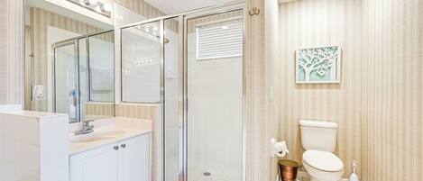 Een moderne badkamer met een glazen douchecabine, een wit wastafeltje met spiegel erboven en een toilet met een decoratief kunstwerk van een boom aan de muur.