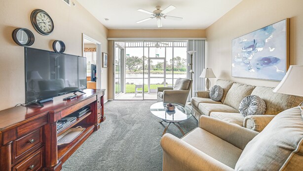 A living room with a flat-screen TV, wooden entertainment center, beige sofa, glass coffee table, and a ceiling fan. - Naples Condo w/ Pond Views & Patio - Near Golf! (Naples)