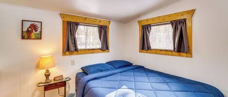 A bedroom with a bed, a nightstand, a lamp, and two windows with curtains.