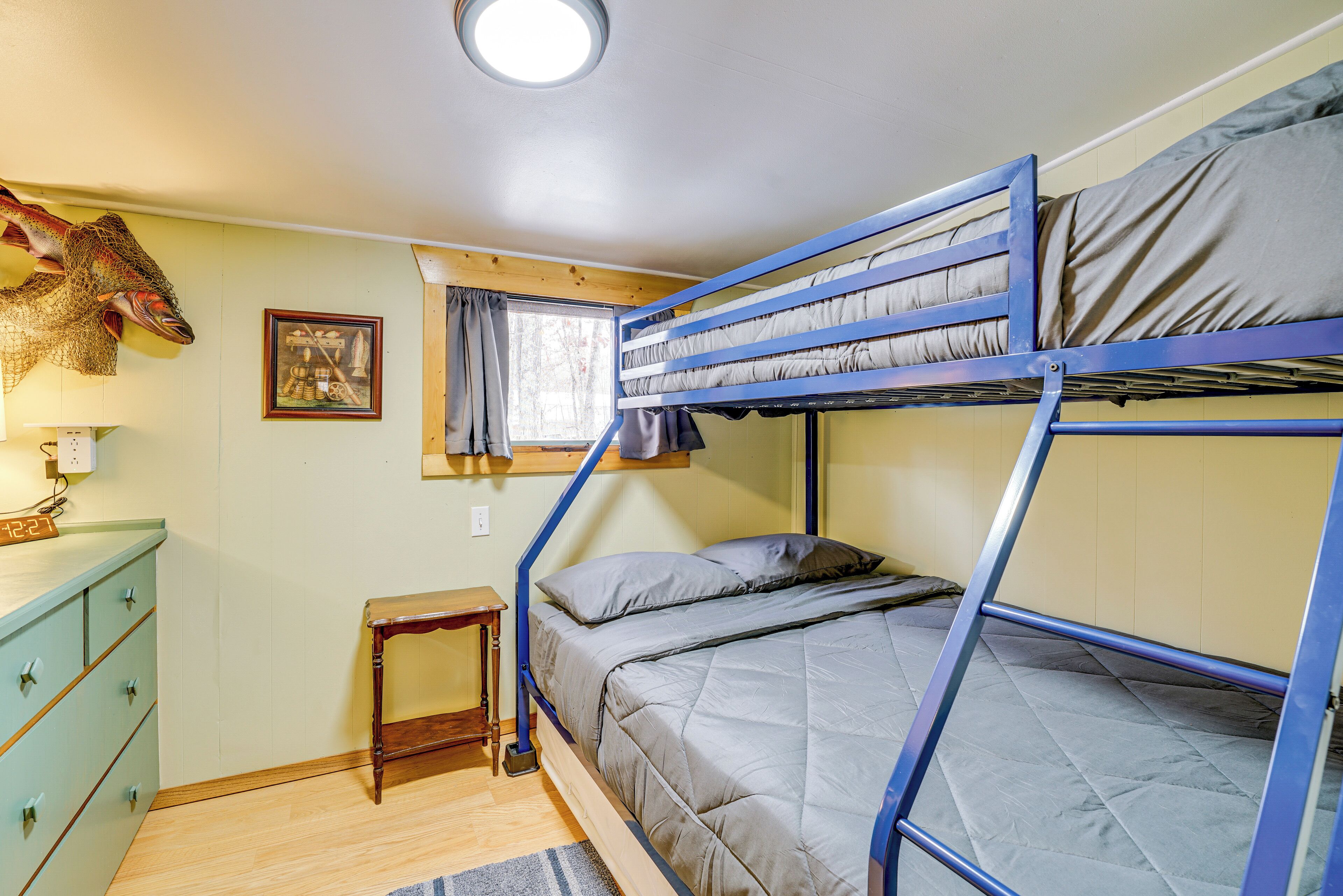 Une chambre avec un lit superposé doté d’une tête de lit en bois, une petite table, une commode et une fenêtre avec des rideaux.