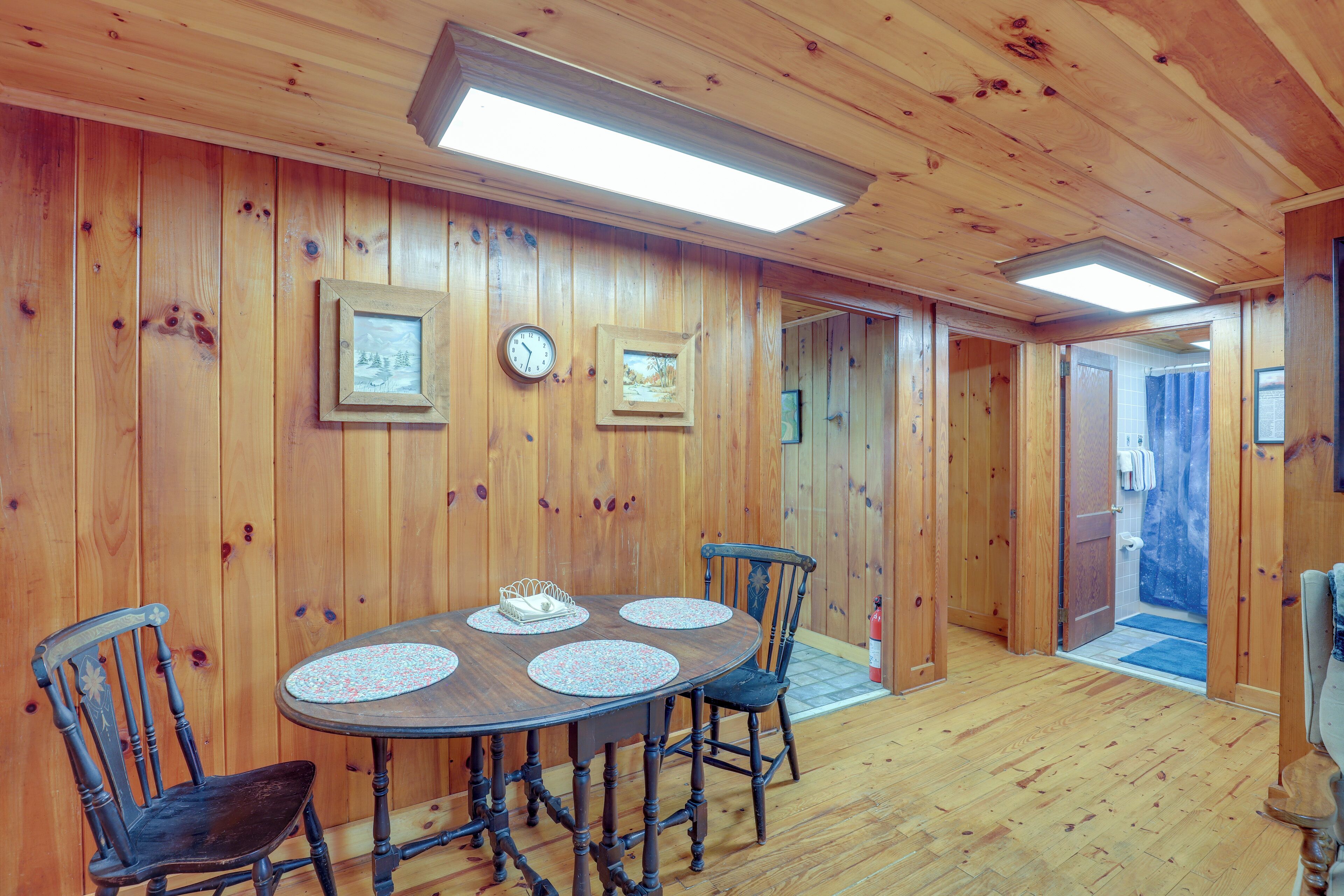 A wooden-paneled room with a round table and four chairs, a clock, and framed pictures on the walls.