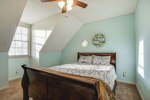 Quarto com teto inclinado, cama de madeira, janela com persianas e espelho fixado na parede.