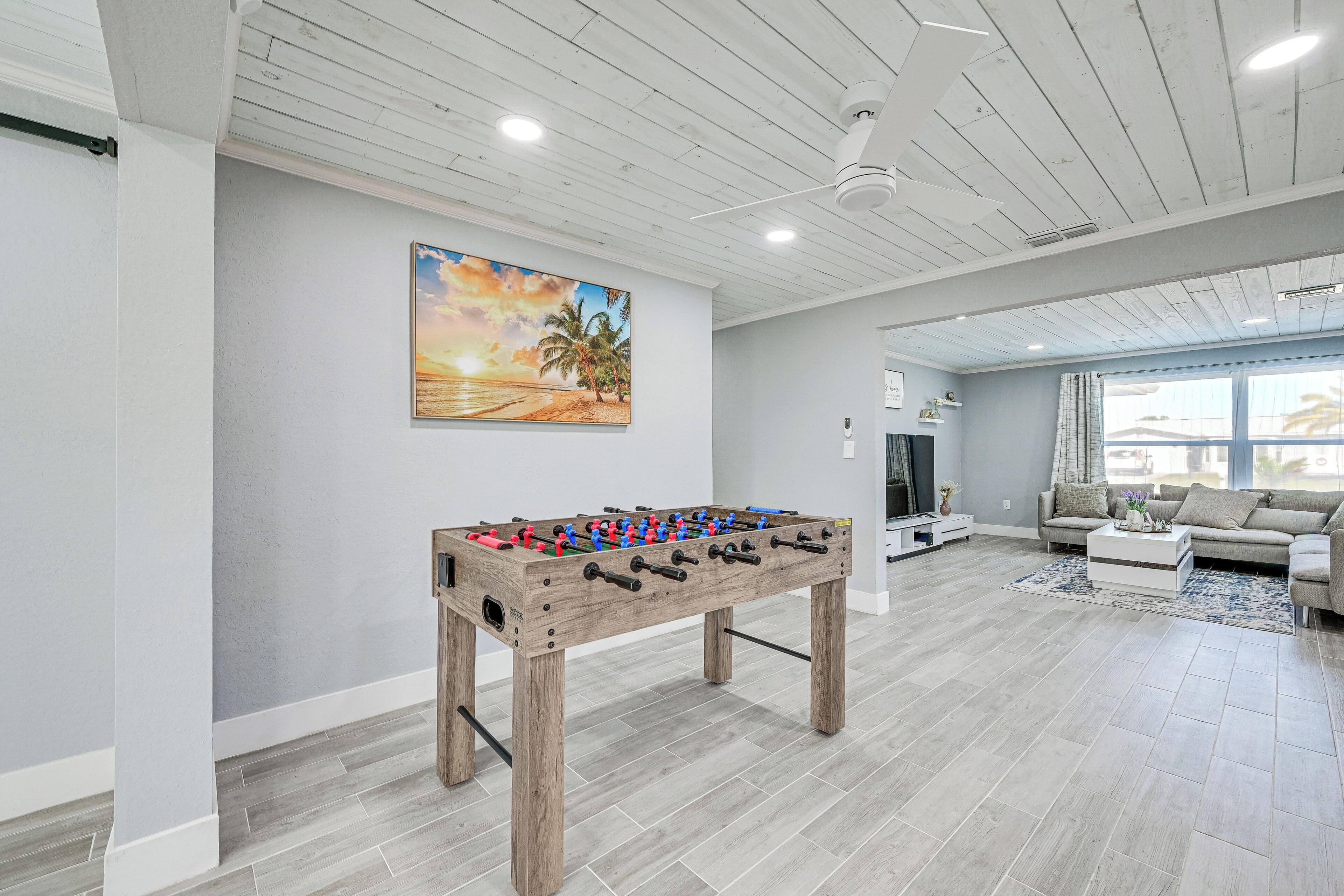 A spacious living room with a wooden foosball table, a large painting of a beach scene, and a modern sofa set.