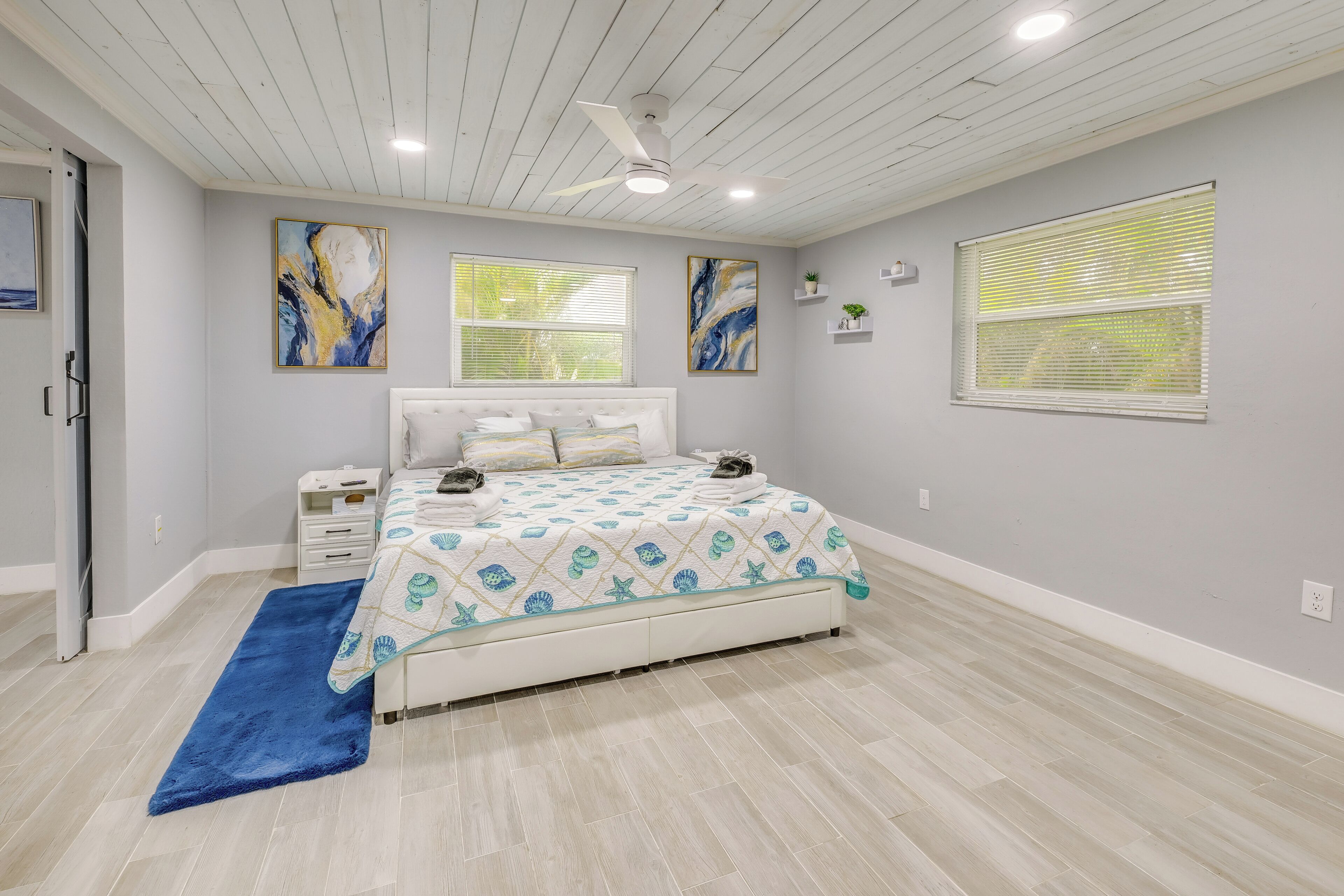Quarto com cama, mesa de cabeceira, ventoinha no teto e duas janelas com persianas.