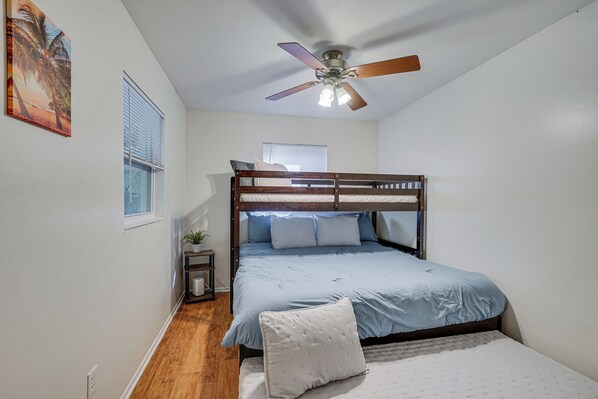 A bedroom with a bunk bed, a ceiling fan, a window with blinds, and a small bedside table with a plant.