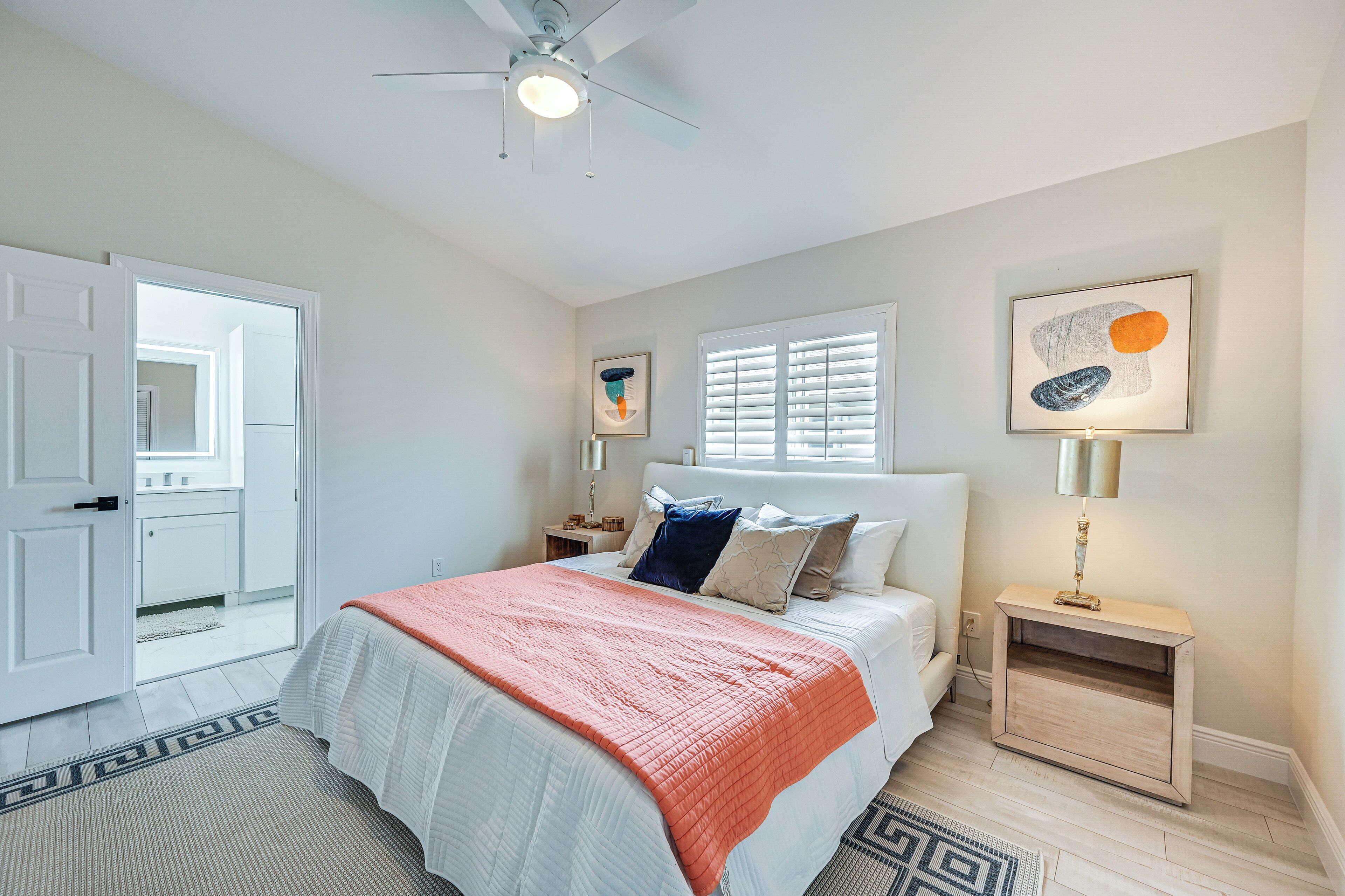 A bedroom with a bed, a nightstand, a ceiling fan, a window with blinds, and a door leading to another room.