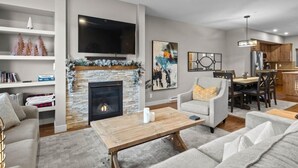 A modern living room with a fireplace, a flat-screen TV, and a dining area with wooden cabinets and a chandelier.