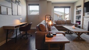 A modern living room with a wooden desk, a sofa, and a coffee table. There are large windows with a view of a house and trees.