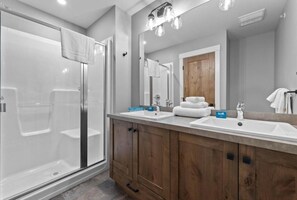 A modern bathroom with a glass shower enclosure, two sinks, wooden cabinets, and a large mirror. - Echo Landing #20 3 Bedroom Townhouse (Sun Peaks)