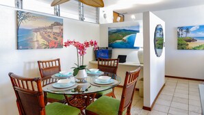 A dining area with a glass table, wicker chairs, and a vase with flowers. There are paintings on the walls depicting tropical scenes.