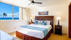 A hotel room with a large bed, two bedside tables with lamps, a ceiling fan, a painting above the bed, and a view of the ocean through a large window.
