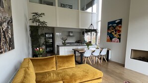 Een moderne woonkamer met een mosterdkleurige bank, een houten eettafel met stoelen, en een keuken op de achtergrond.