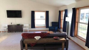 Una sala de estar con un sofá, una mesa de centro, un televisor montado en la pared y un ventanal con cortinas.