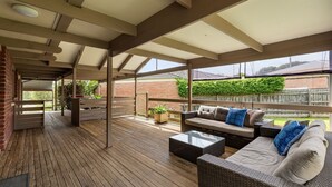 An outdoor patio with wooden flooring, a covered seating area with wicker furniture, and a view of a brick wall and greenery. - Charming Dromana Cottage (Dromana)