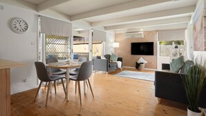 A spacious living area with a wooden floor, a dining table with chairs, a sofa, a TV mounted on the wall, and a potted plant. - Charming Dromana Cottage (Dromana)