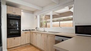 A modern kitchen with wooden cabinets, a built-in oven, a sink, and a window overlooking a brick wall. - Charming Dromana Cottage (Dromana)