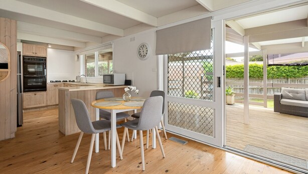 A modern kitchen with a built-in oven, a dining area with a wooden table and chairs, and a sliding glass door leading to a patio with outdoor furniture. - Charming Dromana Cottage (Dromana)