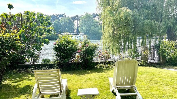 Zwei weiße Loungesessel, die auf einen See blicken, mit einer Weide und einer Steinmauer im Hintergrund.