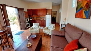 Un soggiorno con un divano marrone, due poltrone, un tavolino in legno e una zona pranzo con tavolo e sedie. Si intravede sullo sfondo una cucina con mobili in legno e un frigorifero.