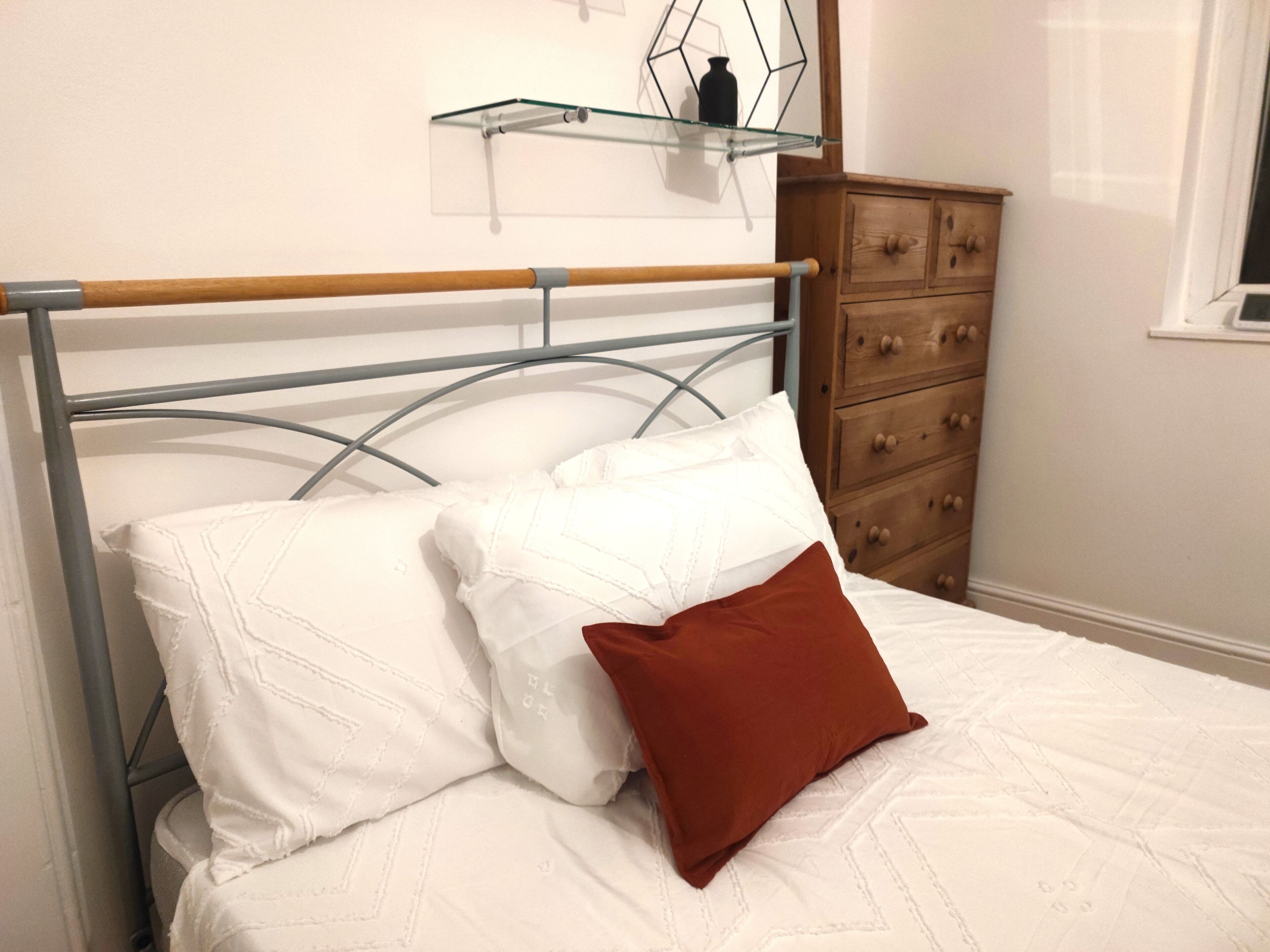 Una cama con estructura metálica, ropa de cama blanca y una almohada marrón. A la derecha hay un aparador de madera y un estante de vidrio encima de la cama.