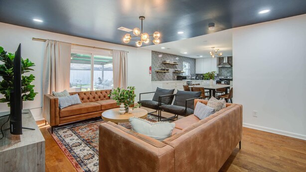 A modern living room with a brown sofa set, a coffee table, and a dining area with a kitchen in the background. - 4-BD Mid-Modern Century with Firepit & Playground (Houston)
