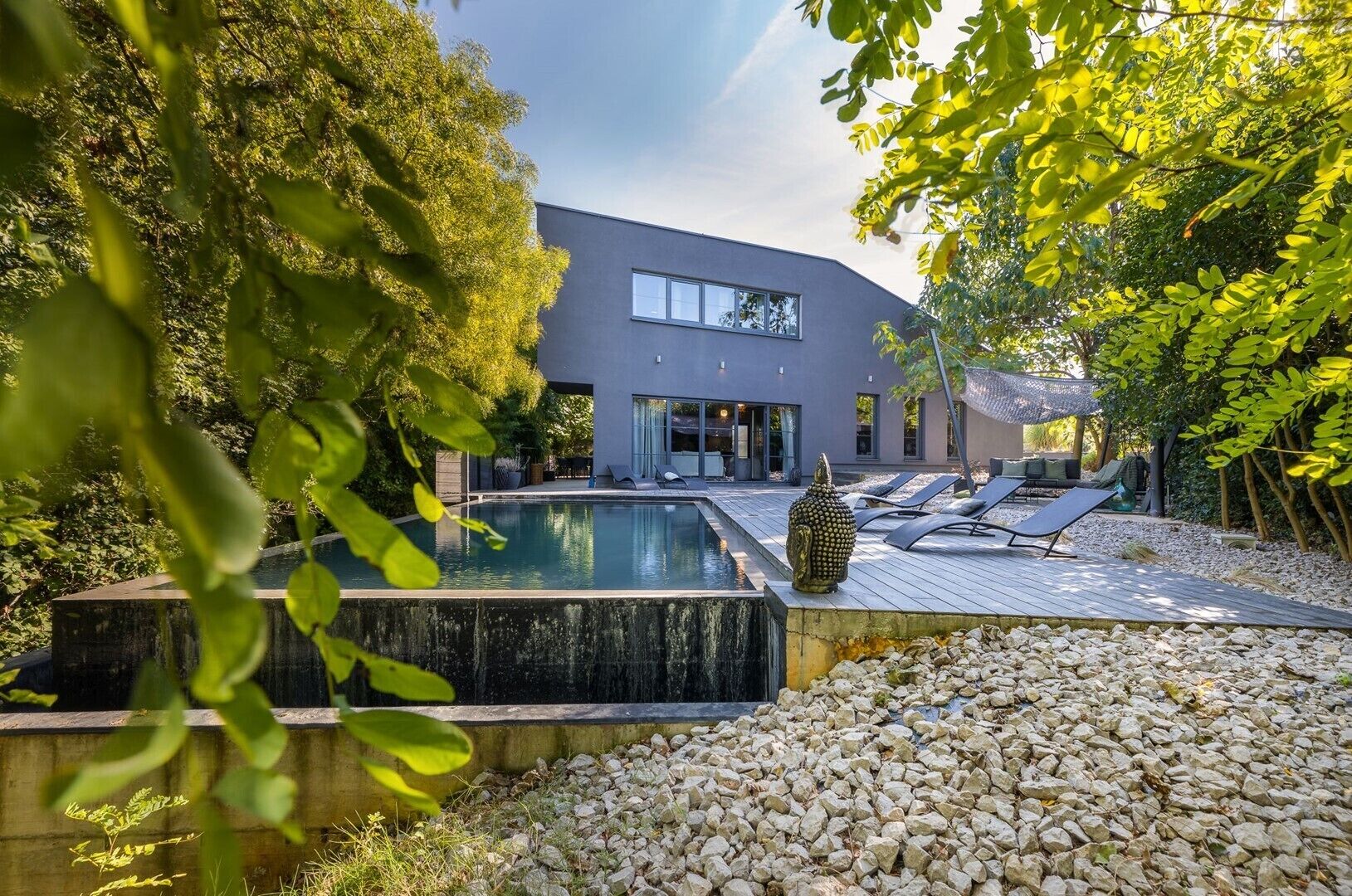 Une maison moderne avec une piscine, entourée d’arbres et dotée d’une terrasse avec des chaises longues.