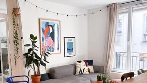 Un salon moderne avec un canapé gris, des chaises en bois, une petite table ronde et une table à manger. Des plantes en pot et des œuvres d’art encadrées ornent les murs.