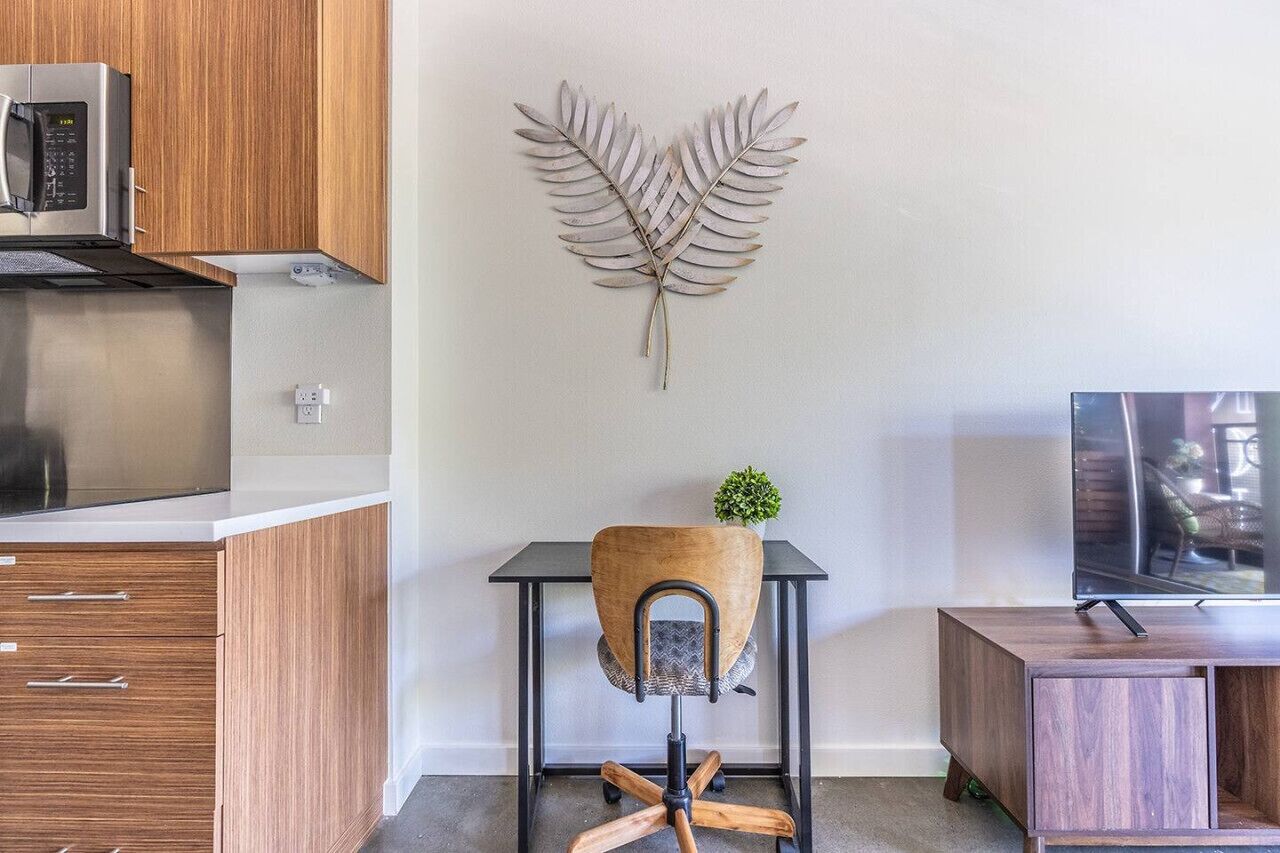 Een moderne keuken met houten kastjes, een RVS magnetron, een decoratie in de vorm van een hart, een kleine potplant, een zwarte bureaustoel en een tv op een houten standaard.