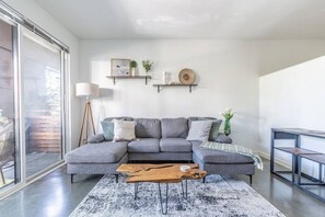 Una sala de estar moderna con un sofá seccional gris, una mesa de centro y una lámpara de pie. Hay estanterías con objetos decorativos y vistas a través de un amplio ventanal corredero.