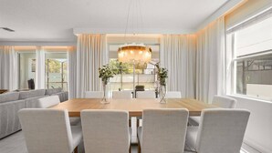 A dining area with a wooden table, six chairs, a chandelier, and a view of the outdoors through large windows.