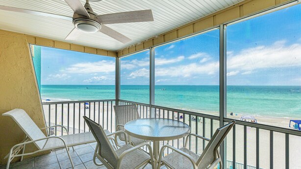 Un balcon avec une table et des chaises en verre, donnant sur une plage aux eaux turquoise et limpides, et sur laquelle on aperçoit un bateau au loin.