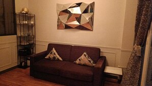 A living room with a dark brown sofa, two patterned throw pillows, a geometric wall art piece, a small side table, and a decorative shelf with glassware. - MARAIS LOVELY BEAUBOURG A (Paris)