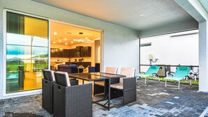 Un espace patio avec une porte vitrée donnant sur la cuisine, un coin salon extérieur avec des meubles en osier, et un espace au bord de la piscine avec des chaises longues.